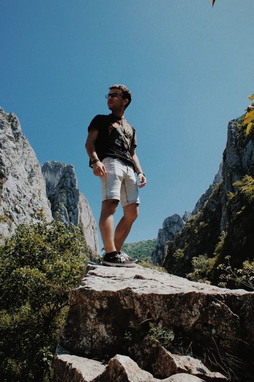 Free A man standing on a rocky cliff surrounded by dramatic mountain scenery under a clear blue sky. Stock Photo