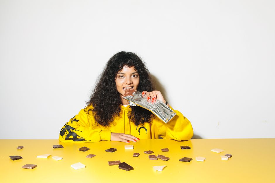A woman in a yellow hoodie delightfully eats a chocolate bar surrounded by scattered chocolate pieces.