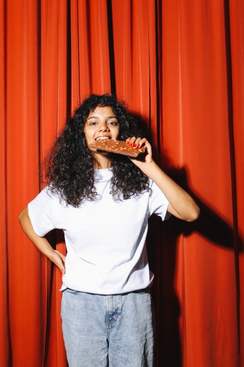 Free Woman with curly hair enjoying a chocolate bar against a vibrant red curtain background. Stock Photo