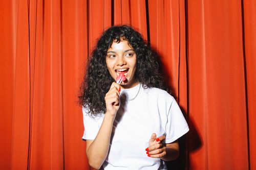 Free Young woman with curly hair in white shirt eating a lollipop against a red curtain. Stock Photo