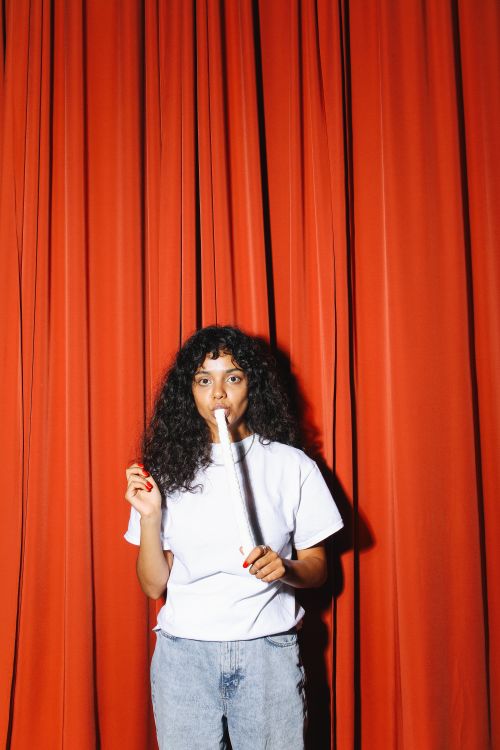 Free Curly-haired woman enjoying a marshmallow on a stick indoors against a vibrant red curtain. Stock Photo