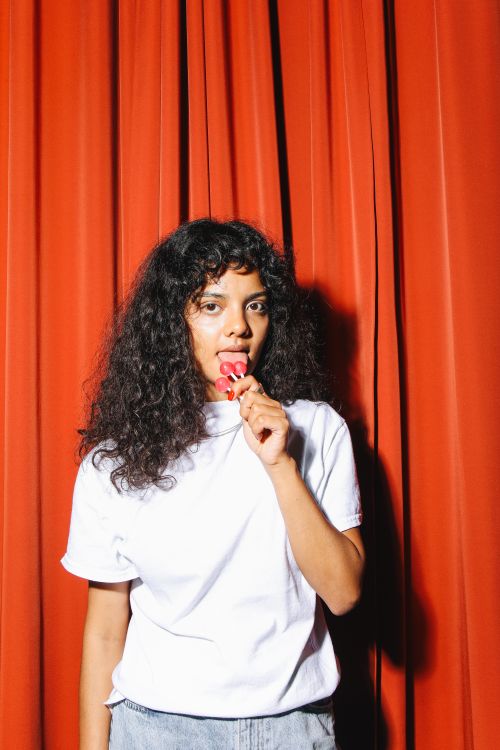 Free Young woman in a white shirt licking a lollipop in front of a red curtain. Stock Photo