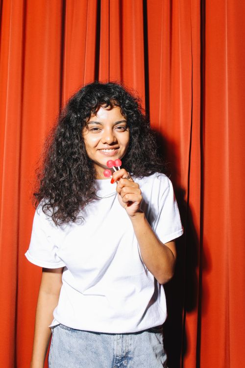 Free A cheerful woman with curly hair holds a lollipop while smiling in front of red curtains. Stock Photo