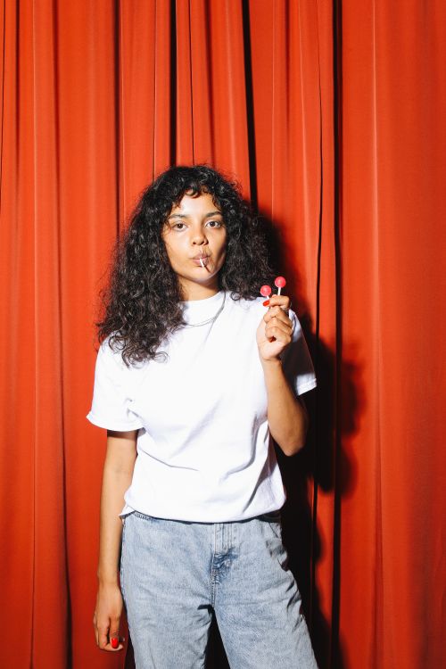 Free A stylish woman poses with a lollipop in front of a red curtain, embodying casual fashion. Stock Photo