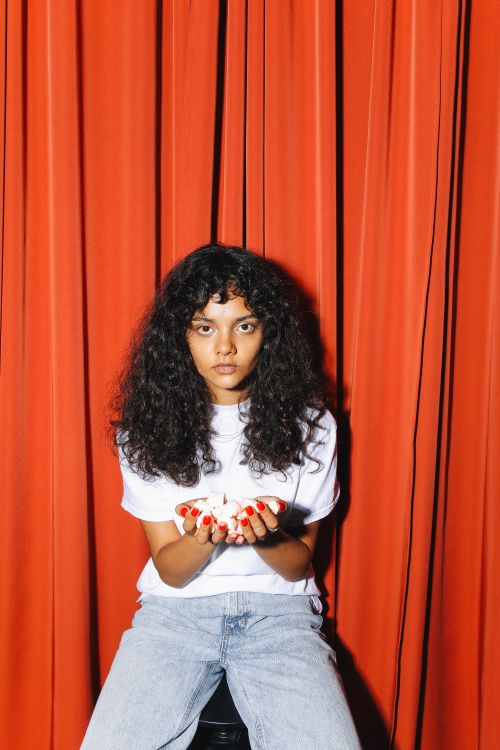 Free Portrait of a young woman with curly hair, sitting against red curtains in a calm and introspective pose. Stock Photo