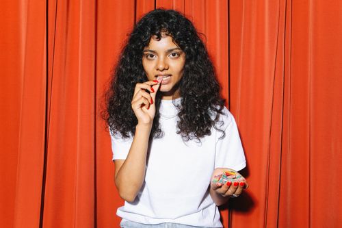 Free Curly-haired woman in white shirt savoring colorful candy against red curtain backdrop. Stock Photo