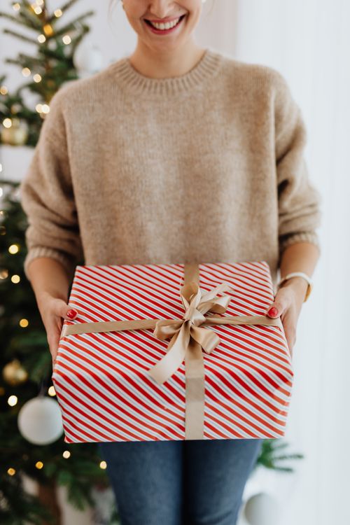 Free Close-up of a woman holding a beautifully wrapped Christmas gift with a tree in the background. Stock Photo