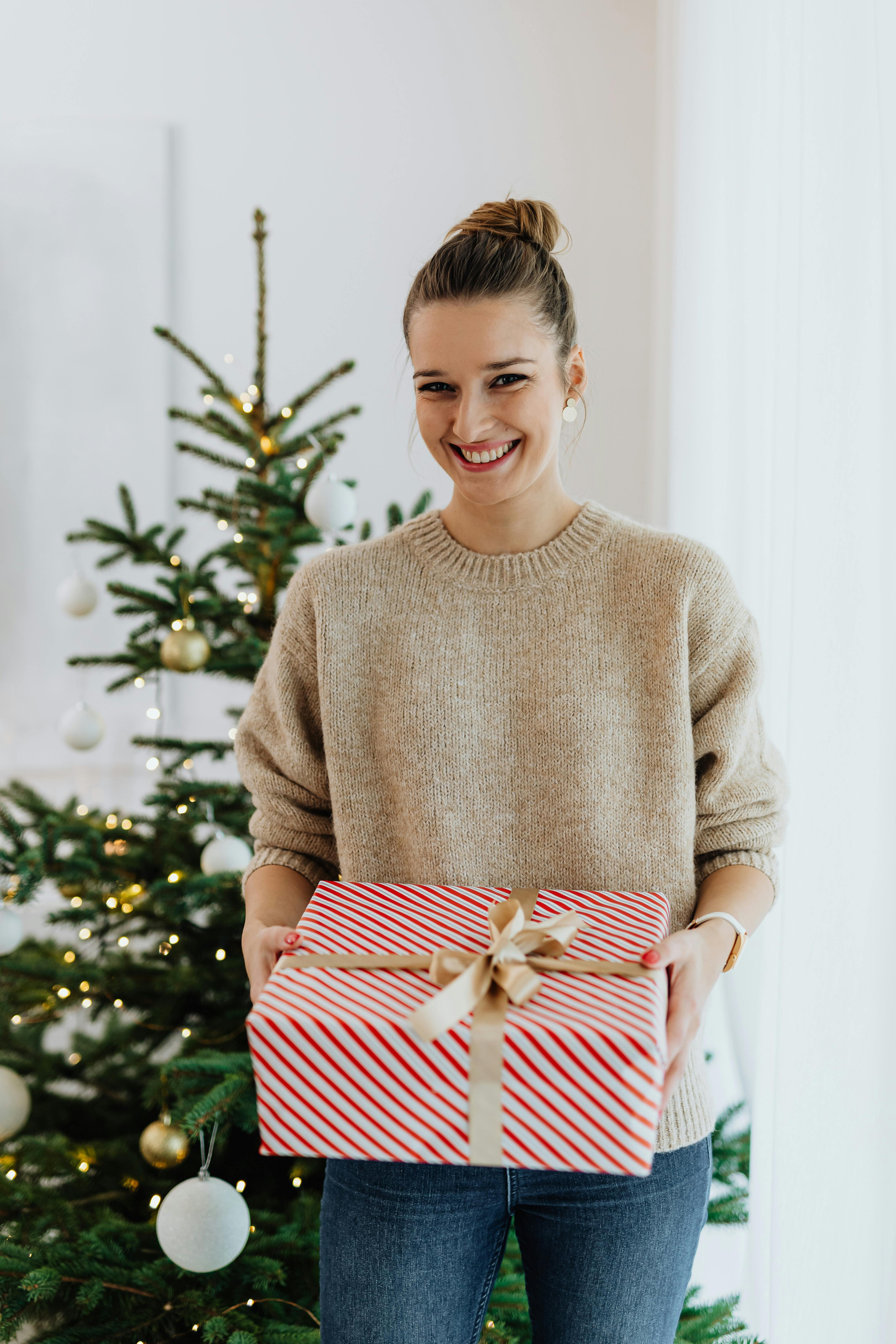 一名妇女穿着舒适的毛衣，在装饰好的室内圣诞树旁拿着一个条纹礼品盒