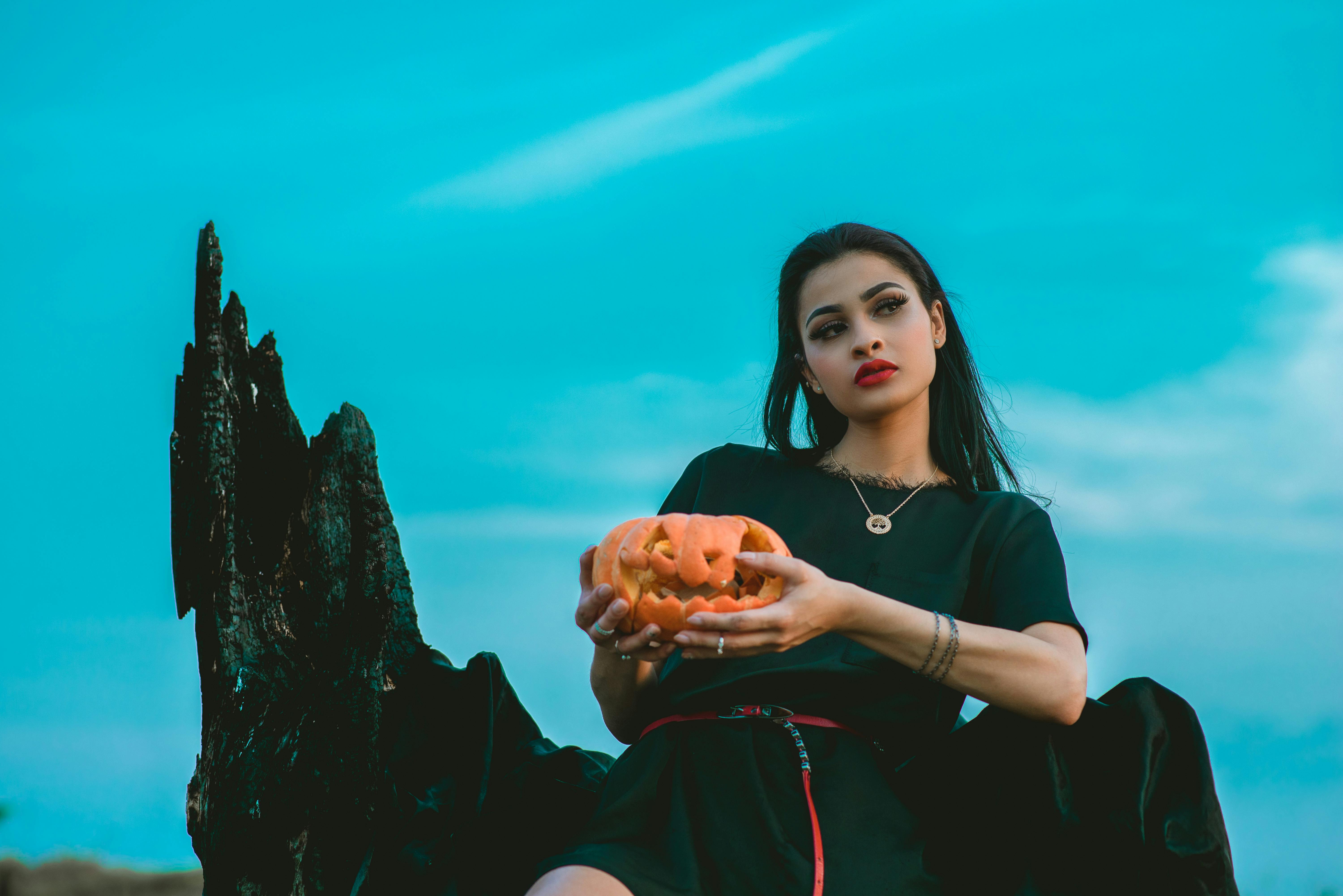 一位黑发时尚的哥特风女子在蓝天下捧着一个雕刻的南瓜