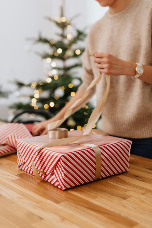Free A woman wraps a Christmas gift with ribbon beside a festive tree indoors. Stock Photo