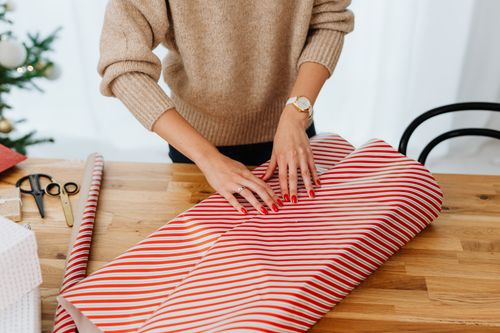 Free A woman wrapping a Christmas gift indoors using red and white striped paper, surrounded by holiday decorations. Stock Photo
