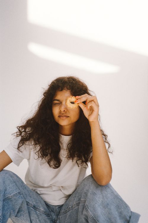 Free A young woman with curly hair poses playfully with a cookie against a bright background. Stock Photo