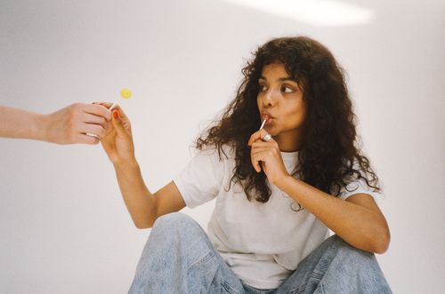 Free Young woman with curly hair in casual attire savoring a lollipop indoors. Stock Photo