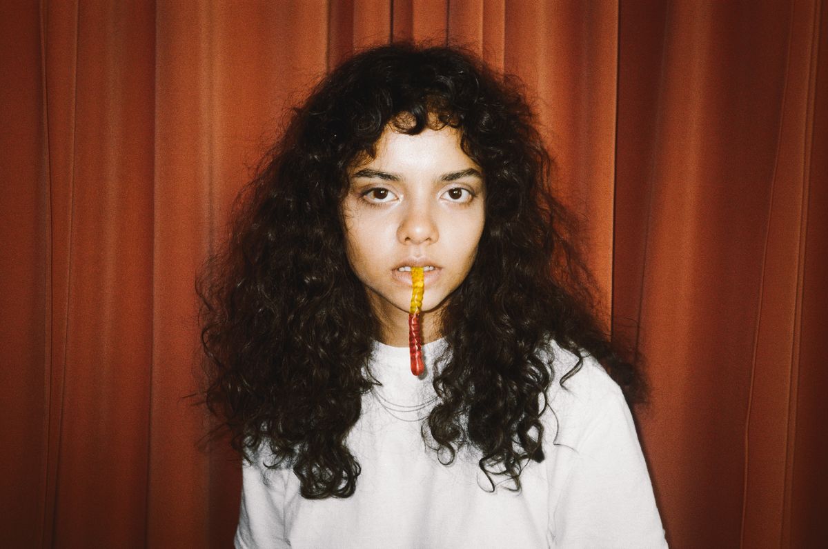 Young woman with curly hair holding a gummy worm against a curtain backdrop.