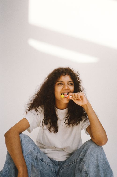 Free Woman with curly hair sitting casually enjoying a lollipop against a bright backdrop. Stock Photo