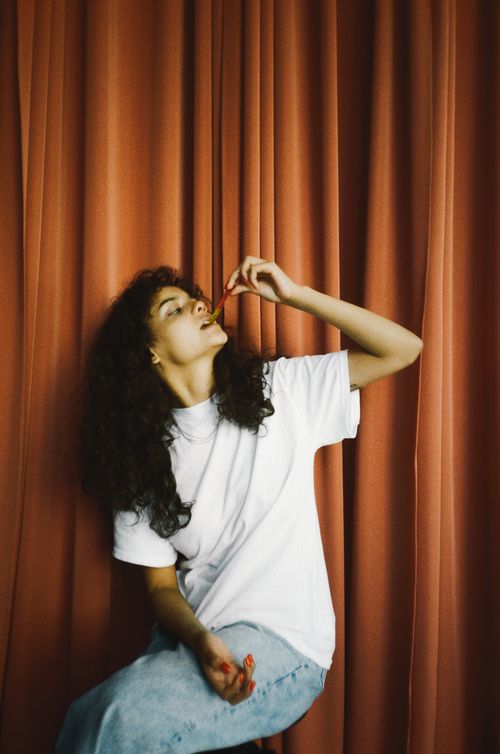 Free A young woman enjoys candy, seated against a vibrant red curtain, capturing a playful and casual moment. Stock Photo