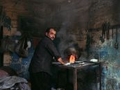 Back view of ethnic worker heating pot with burning flame while looking at camera in smithy