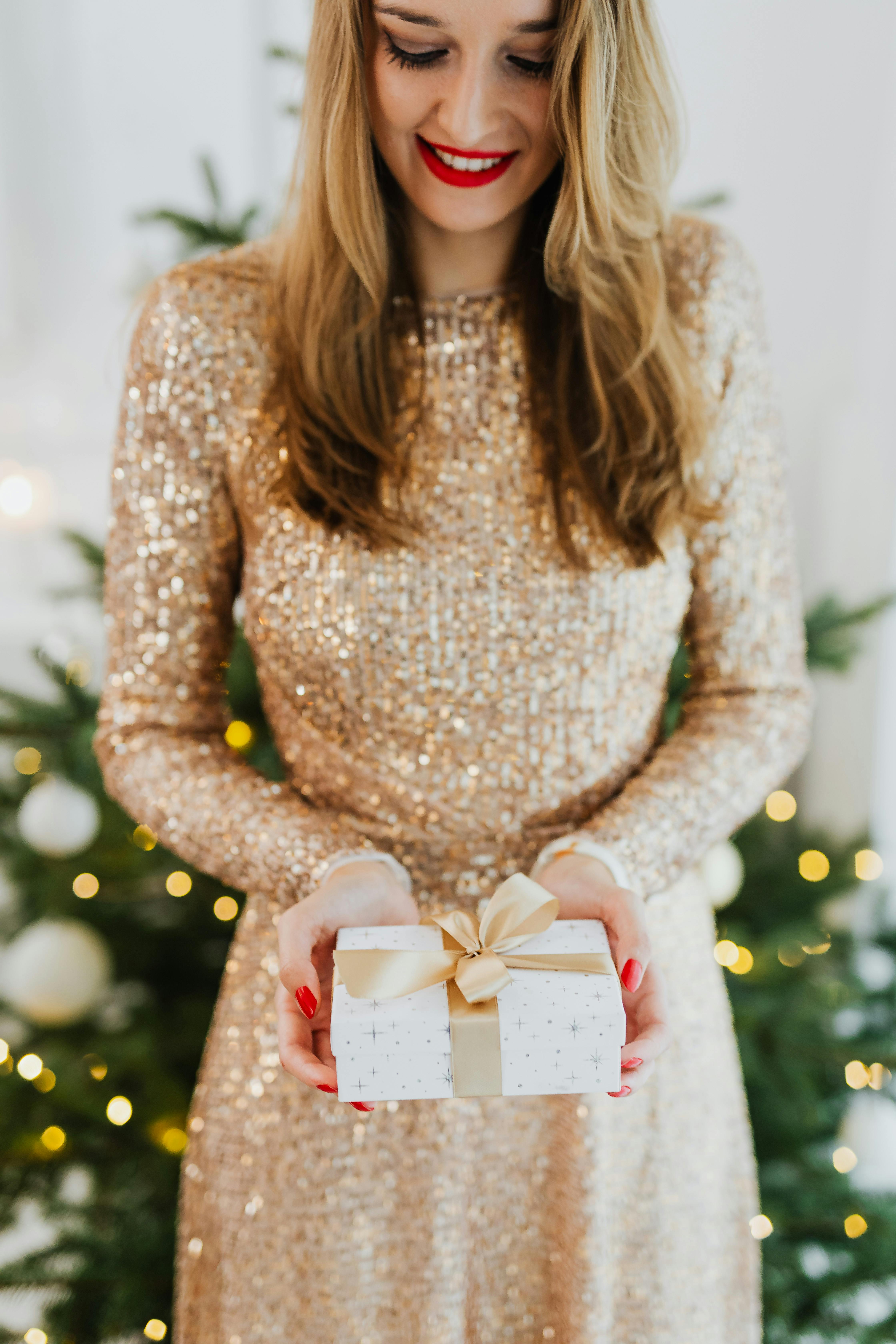 Smiling woman in a sequin dress holding a Christmas gift in front of a decorated tree.