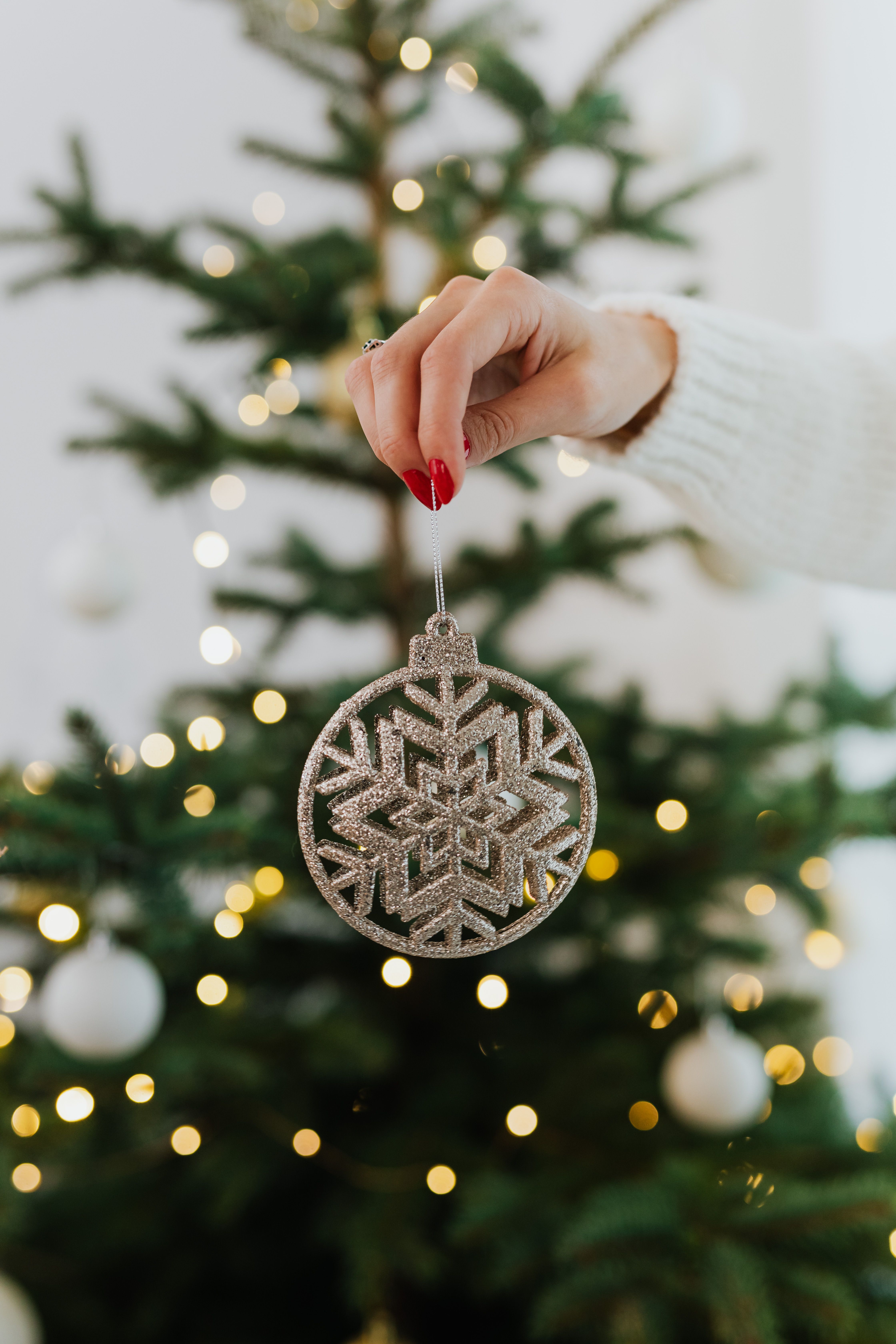 hand with red nails holding a silver snowflake ornament in front of a decorated Christmas tree