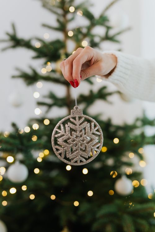 hand with red nails holding a silver snowflake ornament in front of a decorated Christmas tree