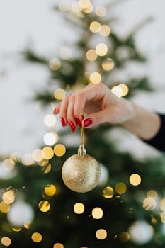 A hand decorates a Christmas tree with a gold ornament, surrounded by festive bokeh lights.