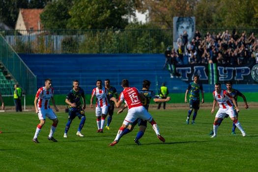 Intense soccer game with teams in action under bright sunlight, surrounded by cheering fans.