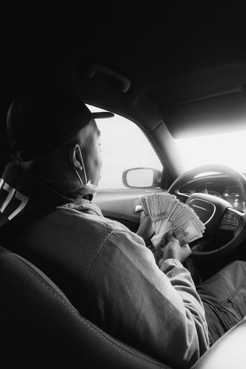 Grayscale Photo of Man Sitting in the Driver Seat Holding Cash Money
