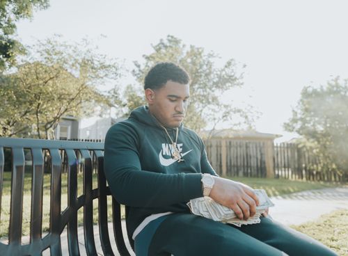 Free Young man in a hoodie counts stacks of cash on a park bench. Stock Photo