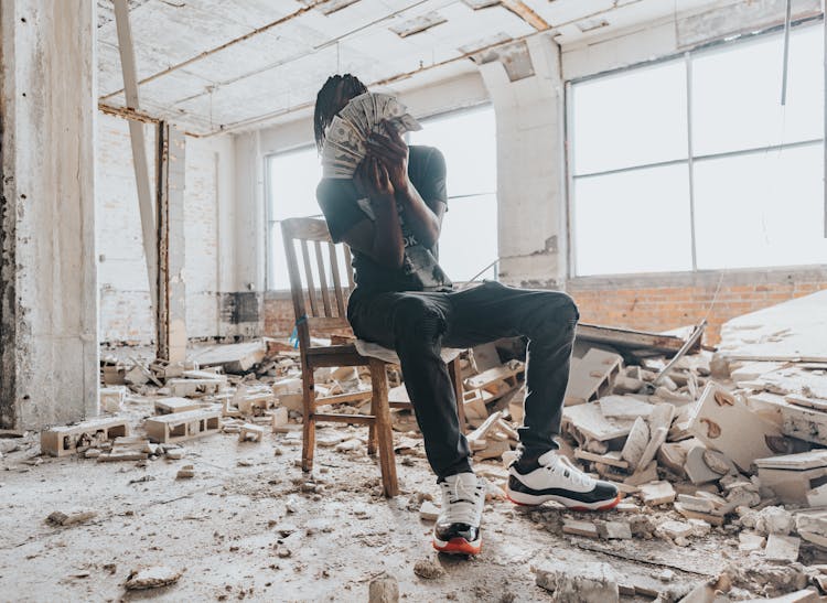 A Person Sitting On A Wooden Chair In A Damaged Building Holding Paper Money