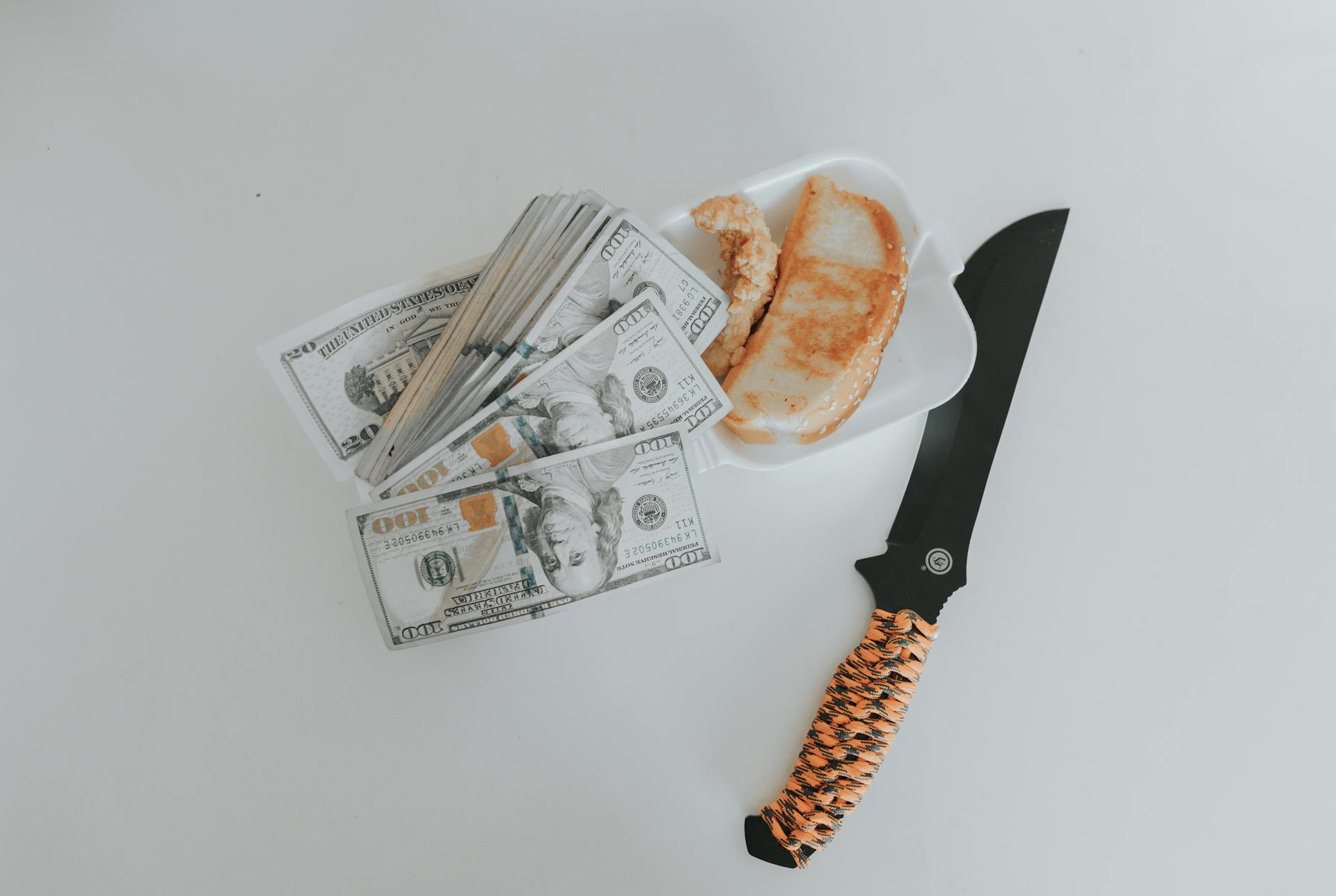 A knife, stacks of hundred-dollar bills, and a food tray on a white background create a compelling still life scene.