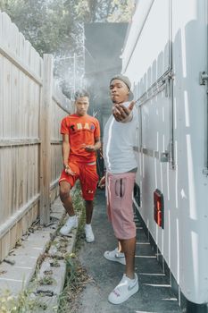Two young men stand and pose confidently in a narrow outdoor alleyway, one is smoking.