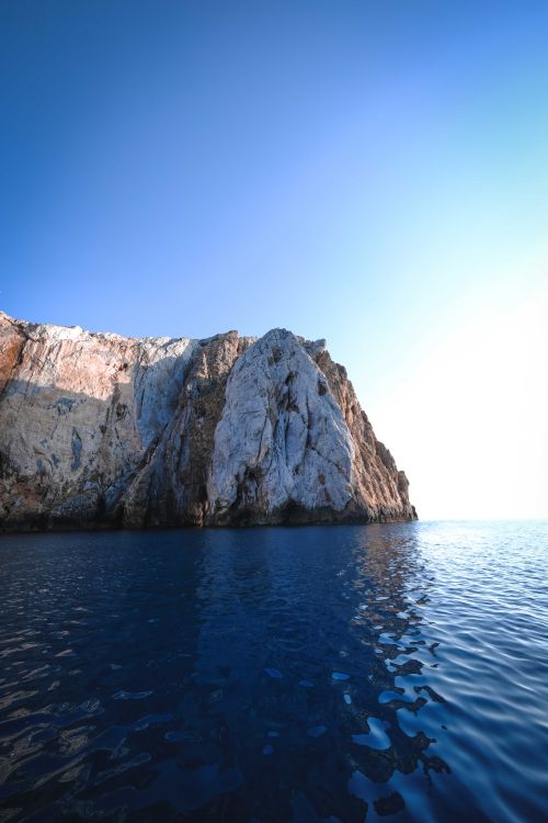 Free Light blue sky over majestic bare steep rock on shore of dark turquoise wavy ocean in sunlight Stock Photo