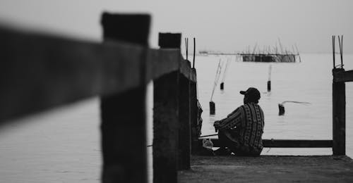 Black and white back view male in casual wear sitting on shabby wooden quay and looking at rippling sea