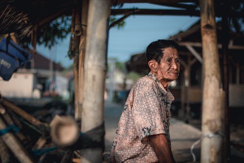 Side view pensive senior Asian male in casual shirt standing in local wooden tent in remote rural village and looking away thoughtfully