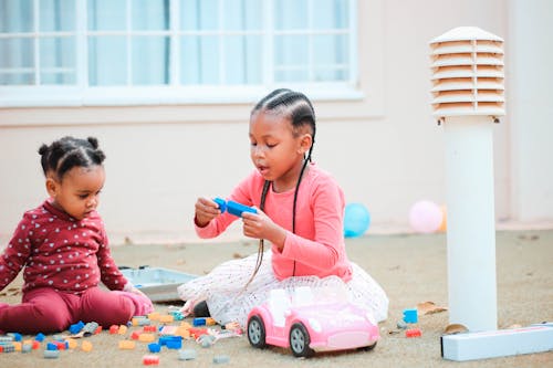 Adorable little African American sisters with stylish hairstyle sitting on floor and plating together with plastic constructor