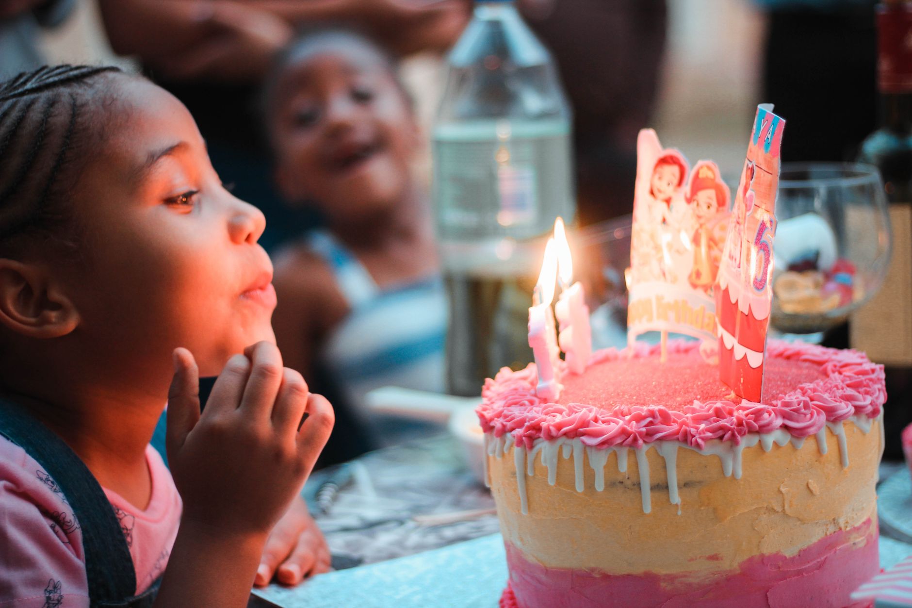 Side view of crop happy little African American girl making wish and blowing out candles during birthday celebration with friends