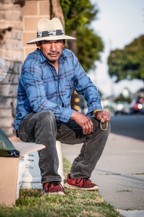 A Man in a Hat and Long Sleeved Shirt Sitting by the Sidewalk