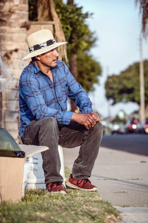 A Man in a Hat and Long Sleeved Shirt Sitting by the Sidewalk