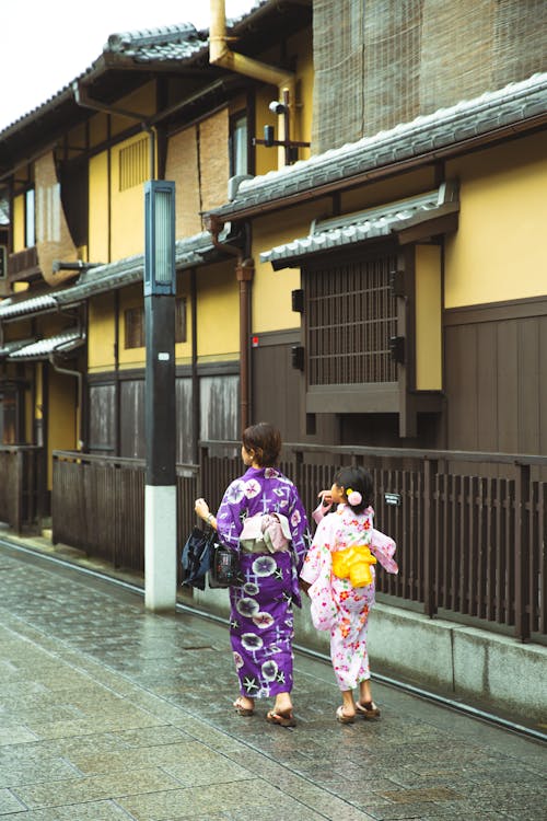 Back view of unrecognizable young Japanese female with little daughter in stylish traditional kimonos walking on street near aged buildings