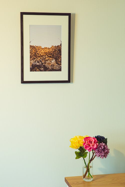 Flores De Colores En Un Florero Sobre Una Mesa De Madera Colocada Cerca De La Pared Con Una Foto Enmarcada