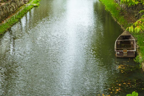 Picturesque calm canal with moored wooden boat rippling along abundant lush greenery in peaceful summer countryside