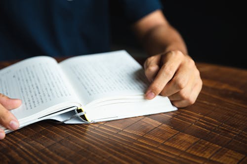 Crop anonymous male wearing dark shirt sitting at round table and enjoying good novel in  hardcover book