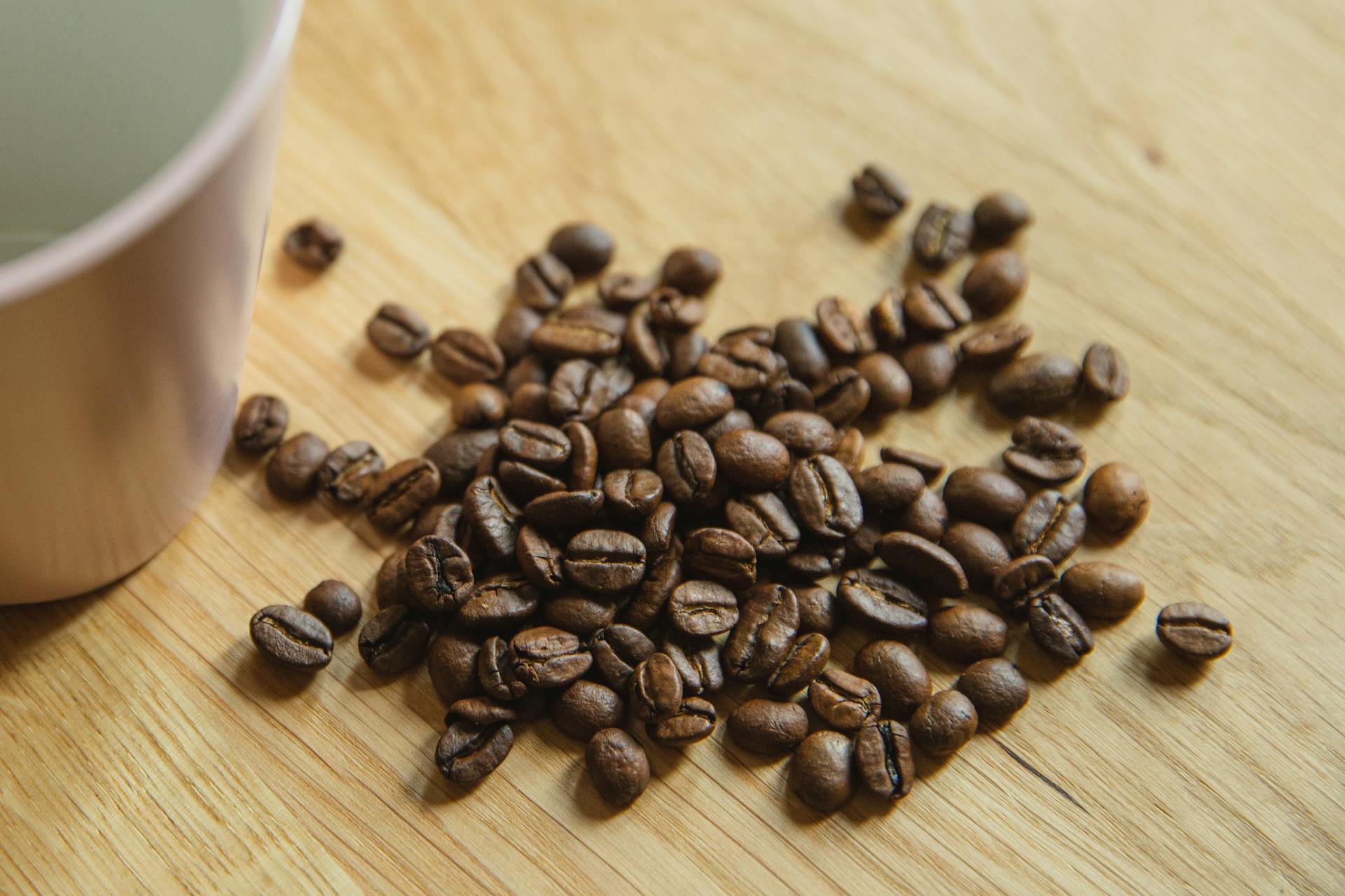 A close-up of aromatic roasted coffee beans next to a pink mug on a wooden surface, exuding warmth and flavor.