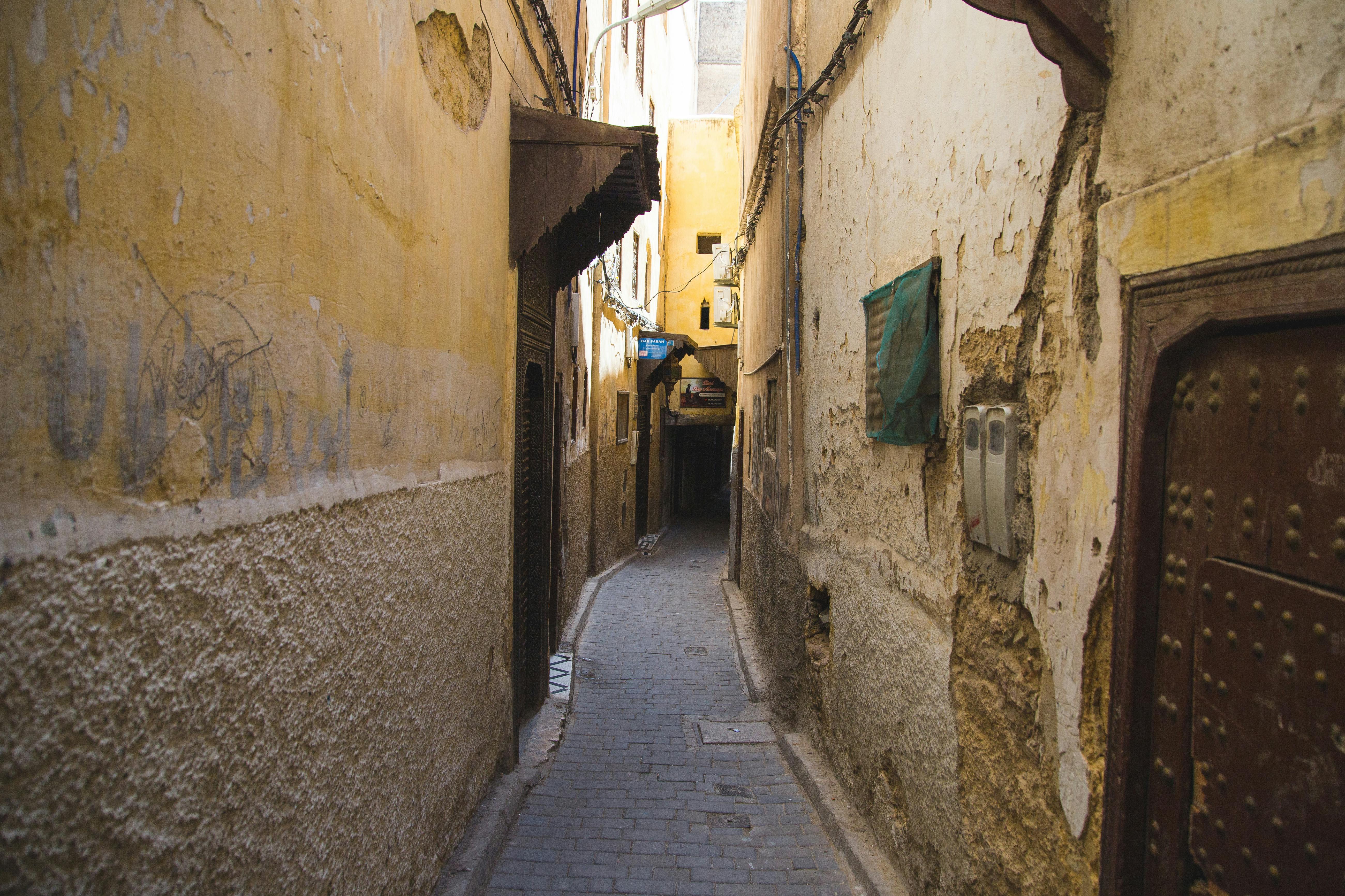 Paved narrow pedestrian walkway between weathered stone residential buildings with metal doors in old Eastern town on sunny weather