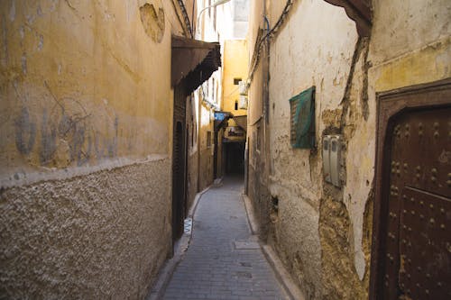 Paved narrow pedestrian walkway between weathered stone residential buildings with metal doors in old Eastern town on sunny weather