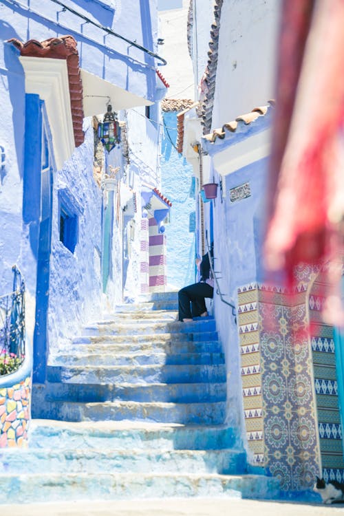 Crop distant person sitting on colorful stairs between blue walls of buildings in sunny street in old town town of Morocco