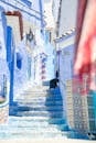 Crop distant person sitting on colorful stairs between blue walls of buildings in sunny street in old town town of Morocco