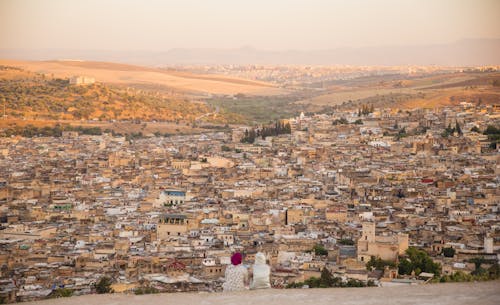 Back view of unrecognizable distant female in traditional wear and headwear sitting on top of mountain above old town with buildings