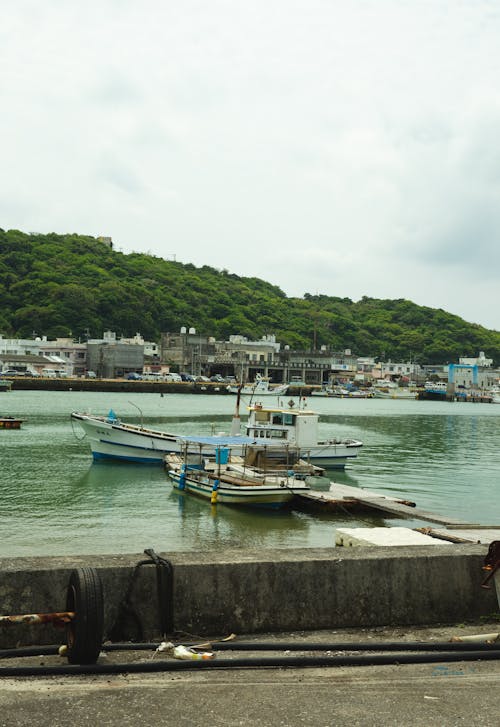 Free Sea vessels floating on rippling water near embankment with stone barrier in coastal town in pier against grassy mountain and cloudy sky Stock Photo