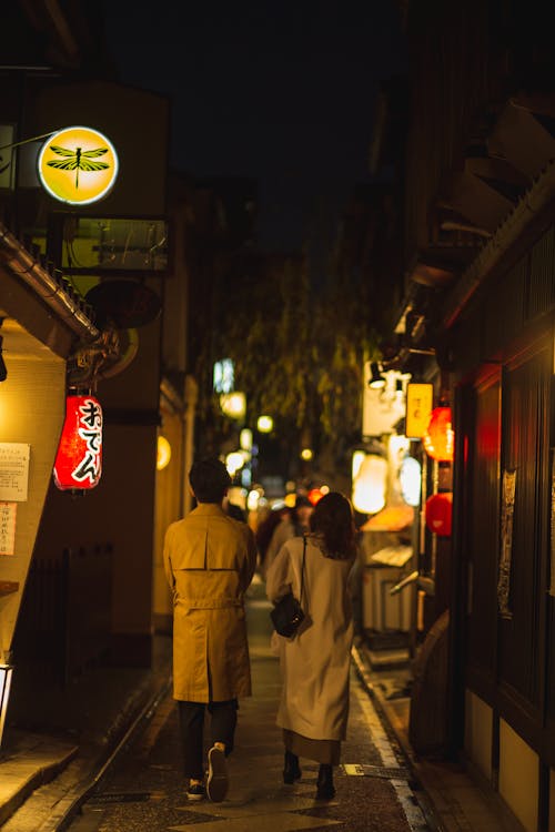 Full body back view of anonymous couple strolling on paved walkway on narrow alley with glowing lanterns in dark city